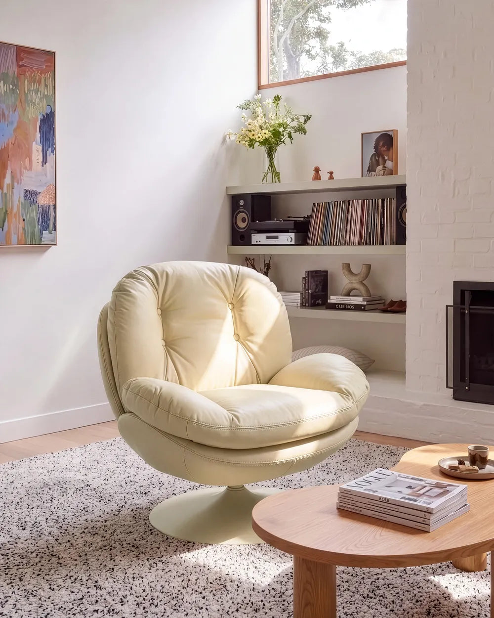 A cream-colored tufted lounge chair in a bright, modern living room setting with a terrazzo rug, wooden coffee table, and built-in shelves.