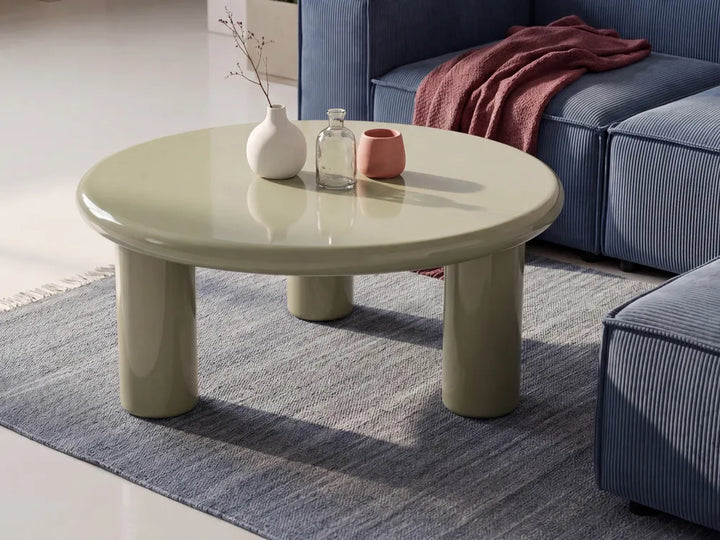 A round glossy low coffee table in a pale sage green color, styled in a living room setting with a blue sofa, a blue rug, and decorative objects on top.