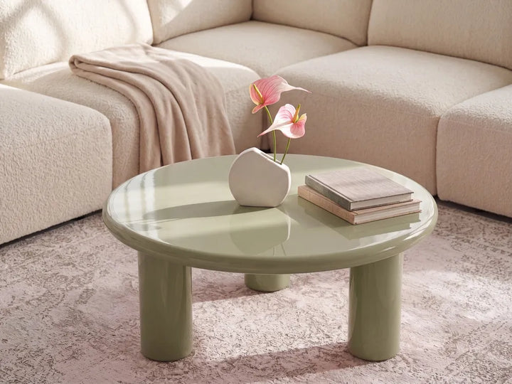 A round low coffee table in a glossy sage green finish, styled in a living room setting with a beige sectional sofa, a pink rug, a white vase with pink flowers, and a stack of books.
