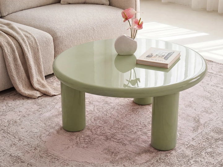 A round low coffee table with a glossy pale green finish and chunky cylindrical legs, styled in a bright living room with a beige sofa, pink rug, and small vase of flowers on top.