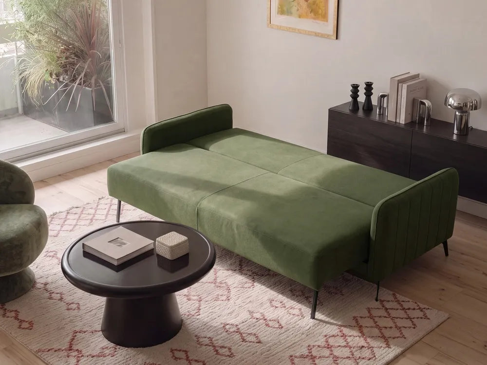 An olive green velvet chaise lounge sofa bed with slim black metal legs, styled in a modern living room setting on a patterned rug next to a round black coffee table.
