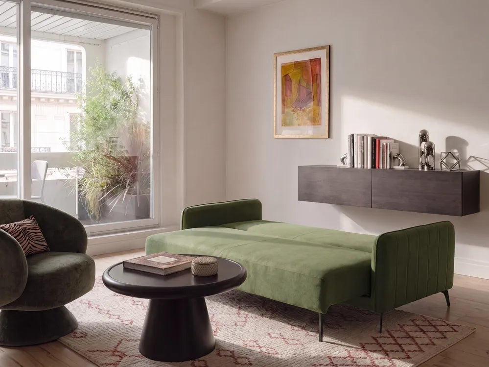 A modern living room featuring a green velvet chaise lounge with slim black legs, a matching green armchair, a round black coffee table, and a patterned rug. The room has light beige walls, a floating dark gray wall shelf with books and decor, and a large window looking out to a balcony.