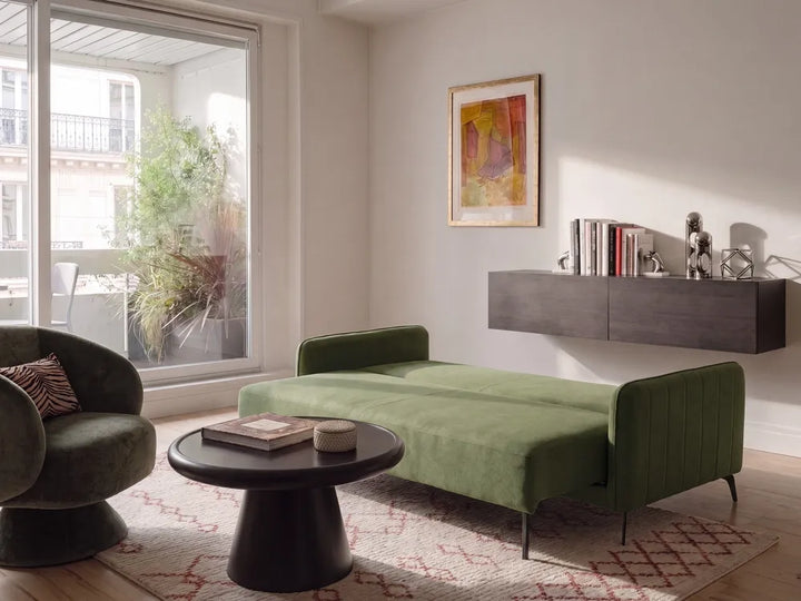 A modern living room featuring a green velvet chaise lounge with slim black legs, a matching green armchair, a round black coffee table, and a patterned rug. The room has light beige walls, a floating dark gray wall shelf with books and decor, and a large window looking out to a balcony.