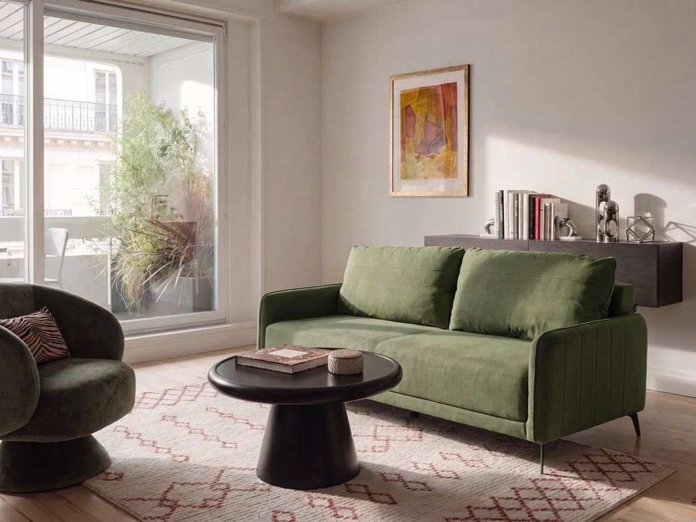 An olive green velvet loveseat sofa in a modern, sunlit living room with a matching accent chair, a black round coffee table, and a patterned rug.