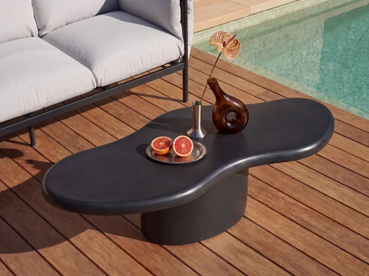 A modern wavy black outdoor coffee table on a wooden deck next to a pool, styled with a glass vase, dried flower, and sliced grapefruit.