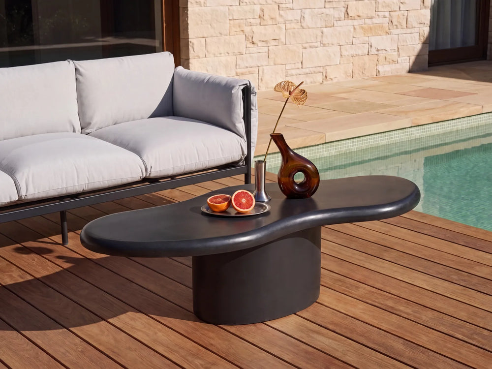 A modern outdoor coffee table with a dark, organic-shaped top and cylindrical base, styled on a wooden deck next to a pool and a light gray sofa.