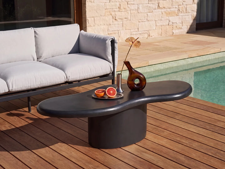 A modern outdoor coffee table with a dark, organic-shaped top and cylindrical base, styled on a wooden deck next to a pool and a light gray sofa.