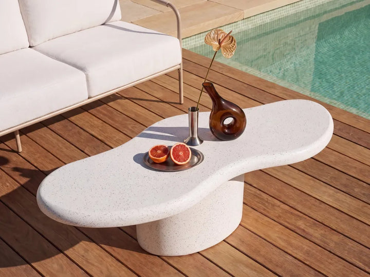 A modern organic-shaped outdoor coffee table in a light speckled stone finish, styled on a wooden deck next to a pool with a white sofa in the background.