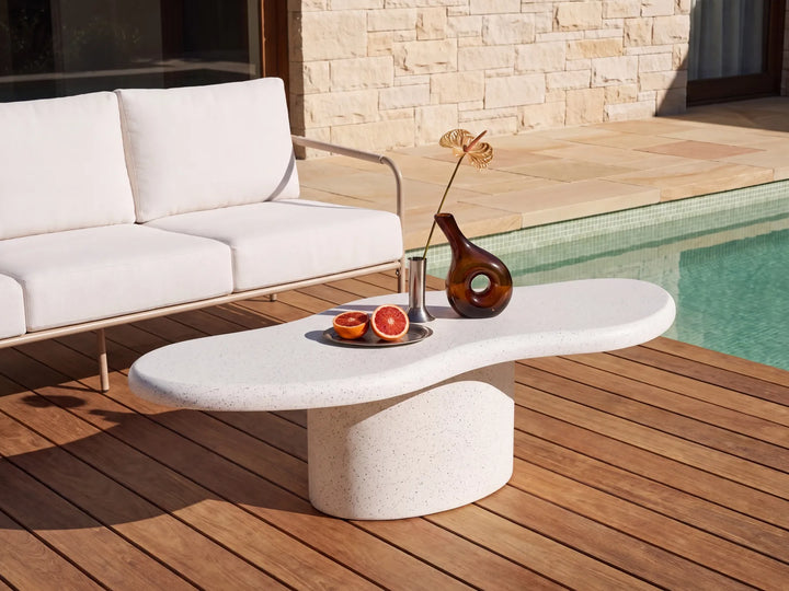 A modern outdoor coffee table with a curved, organic shape and a cylindrical pedestal base, made from a light beige speckled stone. It is styled on a wooden deck next to a white outdoor sofa and a swimming pool, holding a brown glass vase and citrus slices.