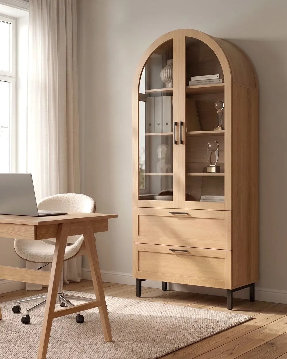 A tall light oak wood arched cabinet with glass doors and two drawers, shown in a modern home office setting next to a wooden desk and chair.