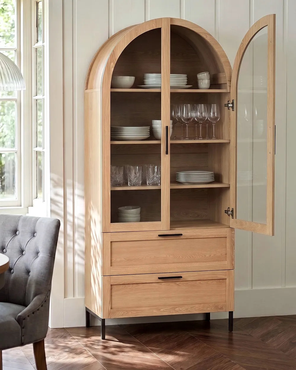 A tall light oak wood hutch cabinet with an arched top, two glass-front upper doors, open shelves with dishes and glassware, two drawers below, and black metal legs, shown in a bright room with a gray tufted chair and window.