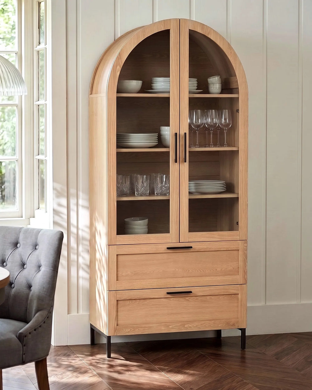 A tall arched wooden display cabinet with glass doors and two drawers, shown in a bright room with a gray tufted chair and wood floor.