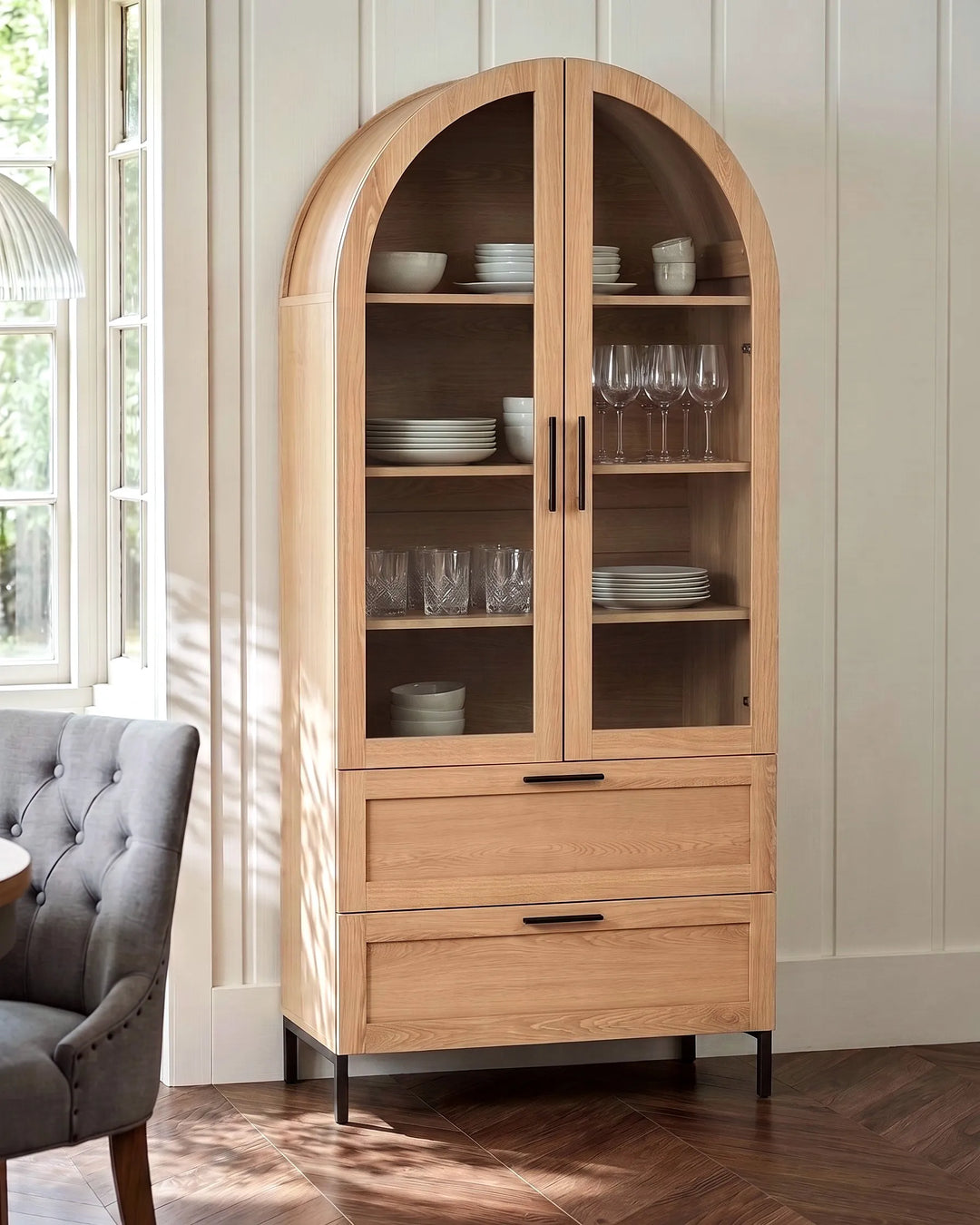 A tall arched wooden display cabinet with glass doors and two drawers, shown in a bright room with a gray tufted chair and wood floor.