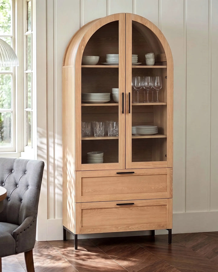 A tall arched wooden display cabinet with glass doors and two drawers, shown in a bright room with a gray tufted chair and wood floor.