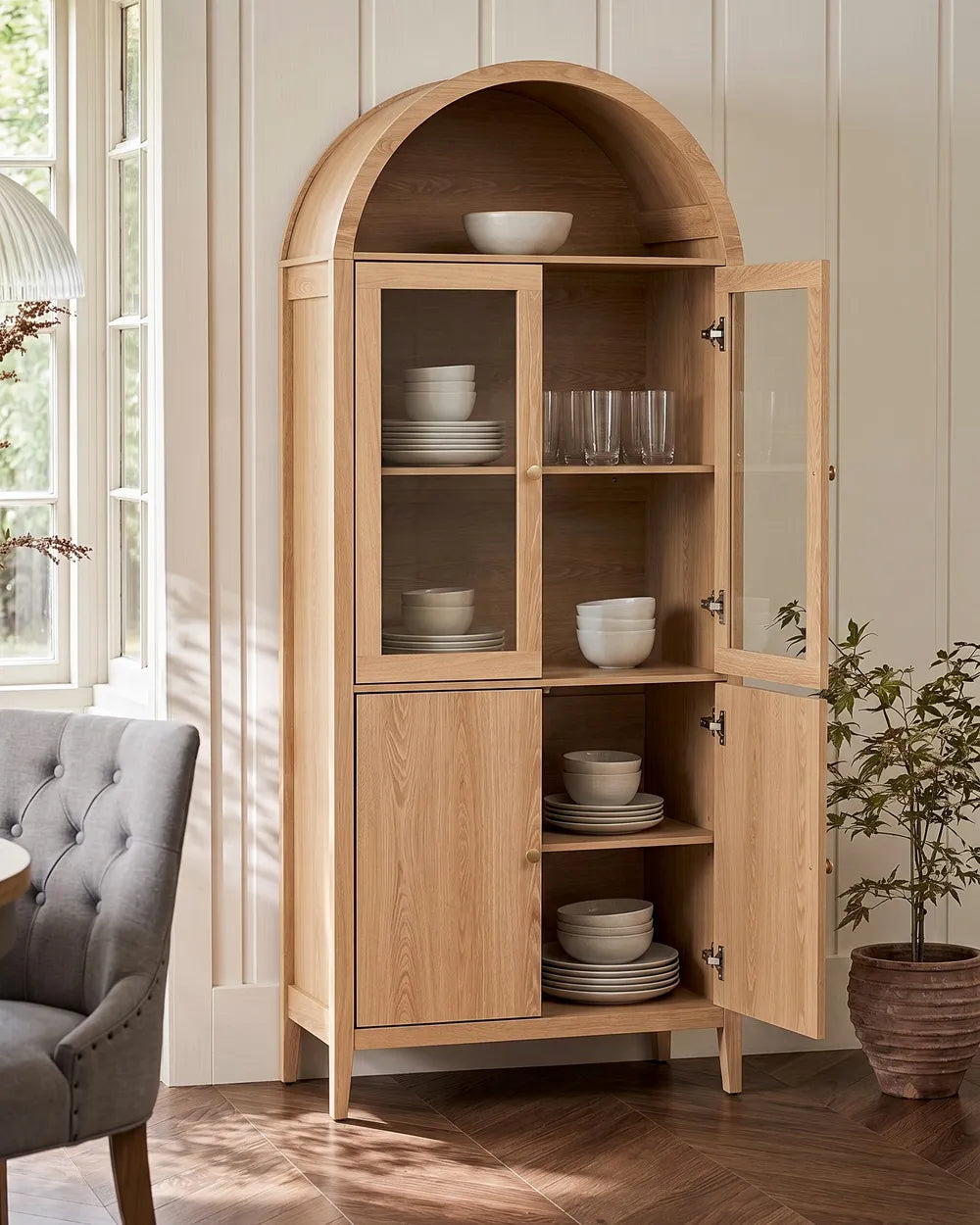 A tall light oak wood arched hutch cabinet with glass doors, shown open and filled with white dishes in a bright room with wood floors and a gray armchair.