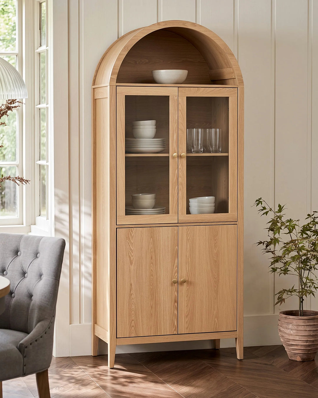 A tall light oak wood hutch cabinet with an arched top, glass-front upper doors, and two lower cabinet doors, shown in a styled room with a gray armchair, window light, and a potted plant.