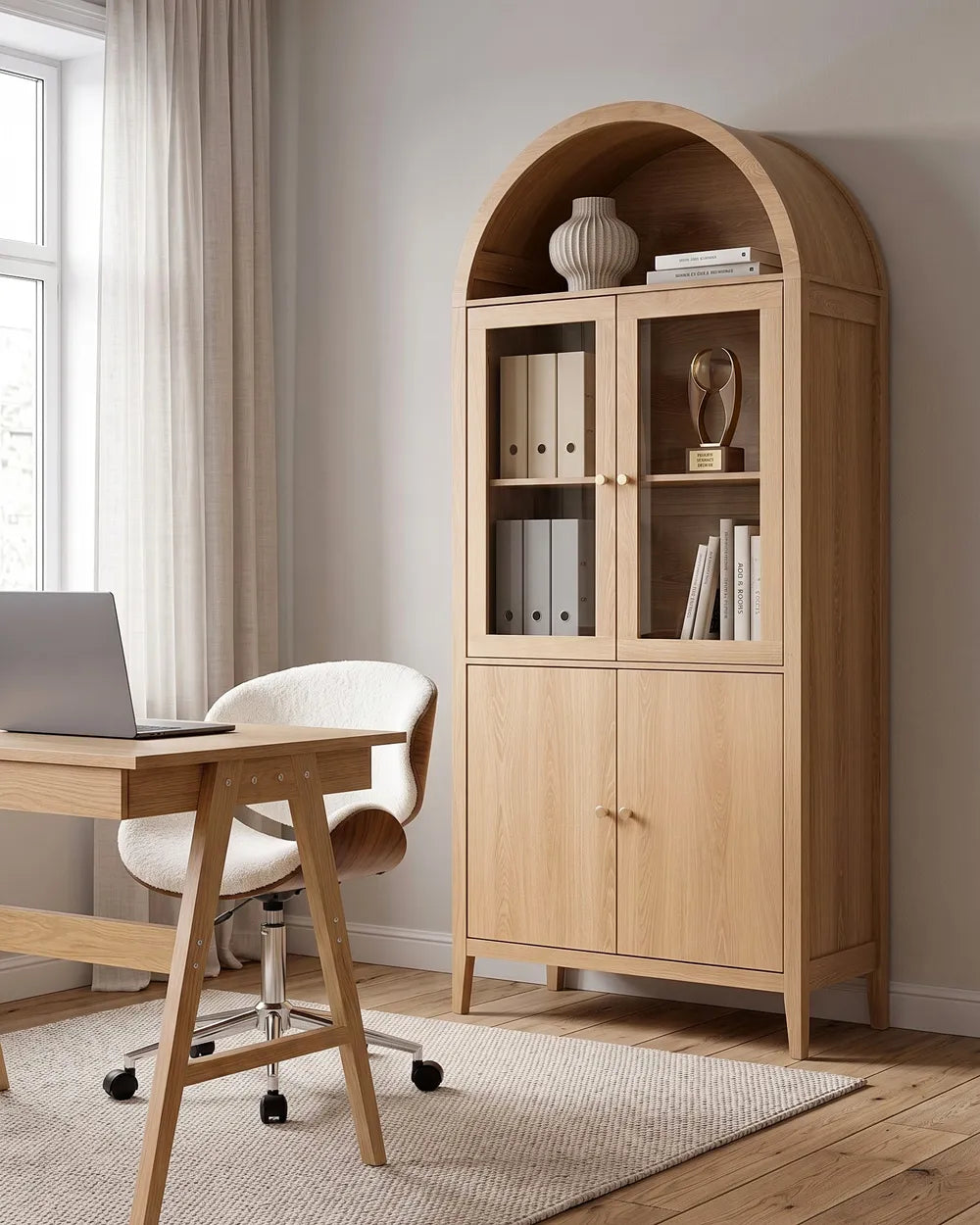 A tall light oak wood arched bookcase cabinet with glass upper doors and two lower doors, styled in a home office next to a wooden desk and chair.