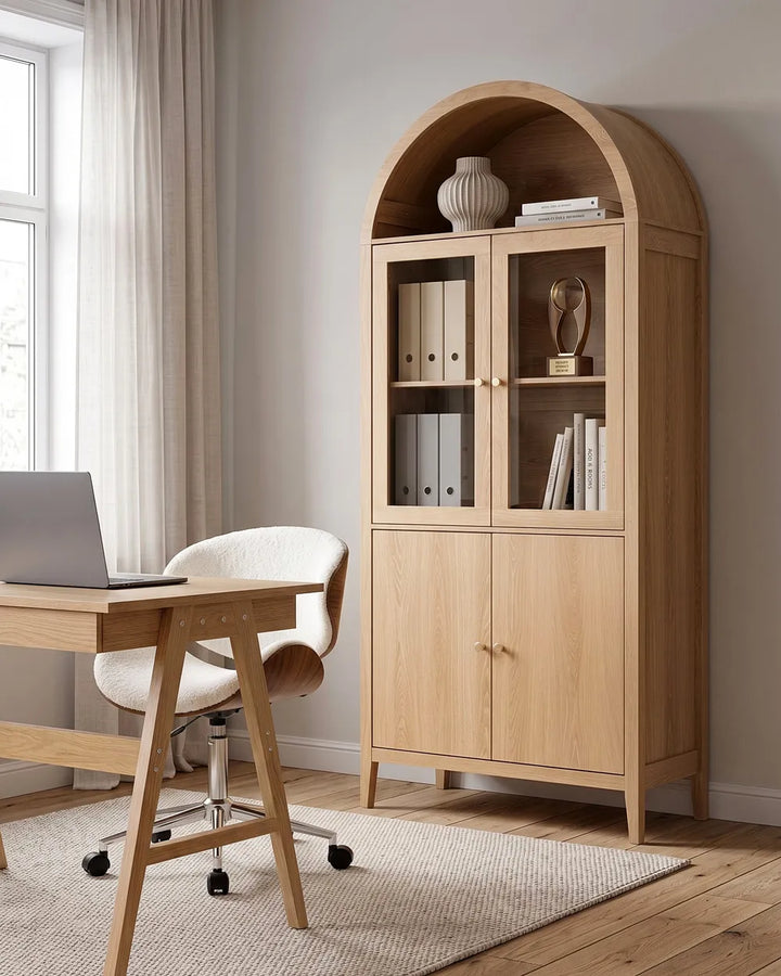 A tall light oak wood arched bookcase cabinet with glass upper doors and two lower doors, styled in a home office next to a wooden desk and chair.