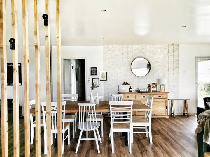 A bright, modern dining room featuring a light wood dining table and white chairs, set in front of a white brick accent wall with a round mirror and a wooden sideboard. The room has hardwood floors and a wooden slat partition in the foreground.