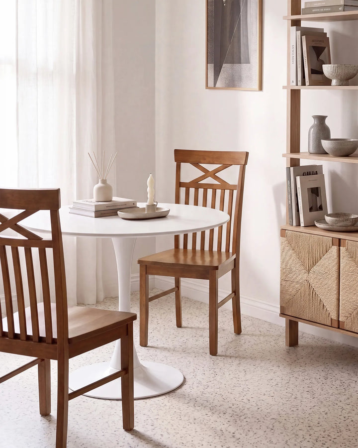 A dining area scene with a round white table and two wooden chairs, plus shelving and decor in a bright interior.