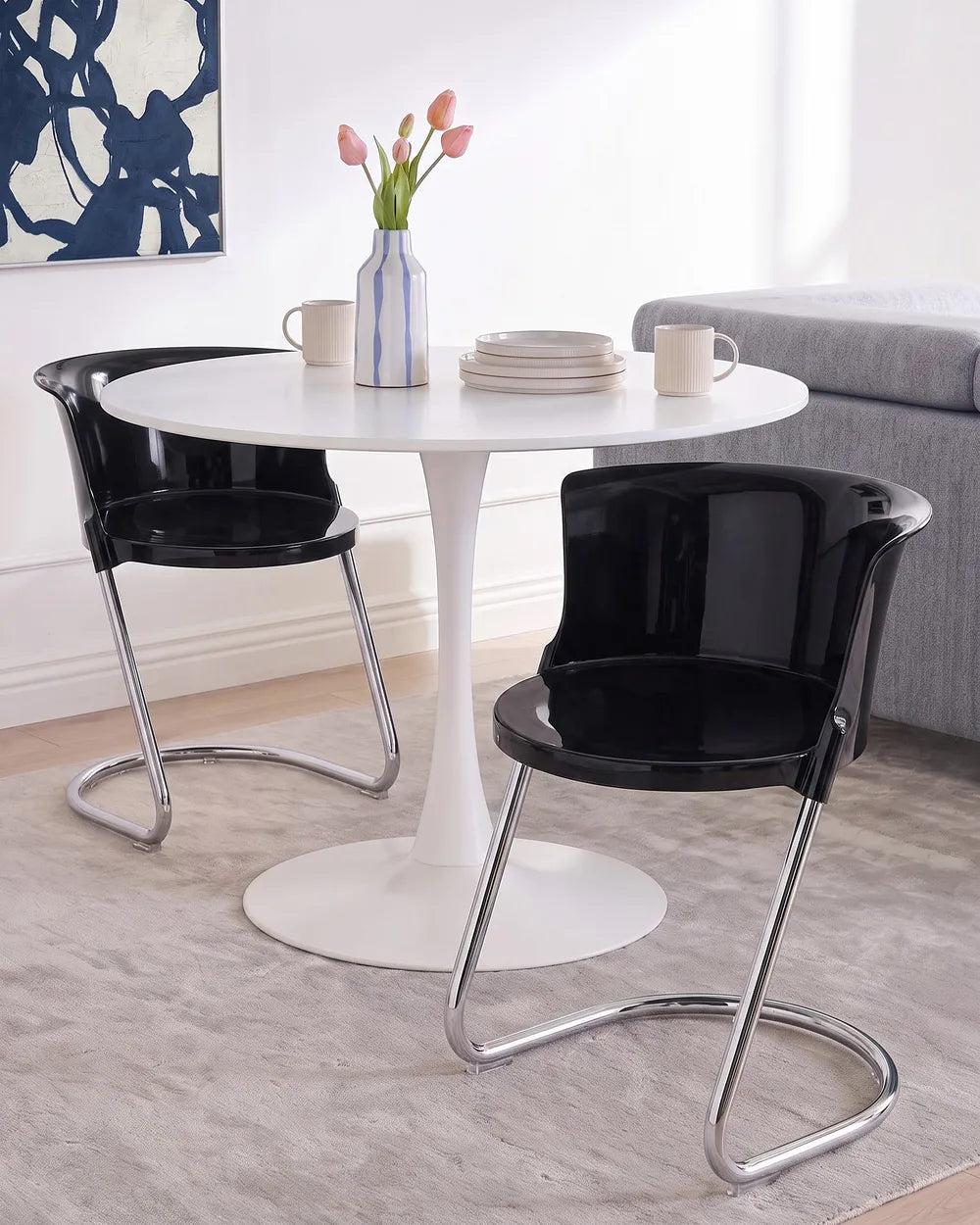 A modern dining setup featuring a round white pedestal table with two black curved-back chairs that have chrome metal legs, set on a light gray rug in a bright room.