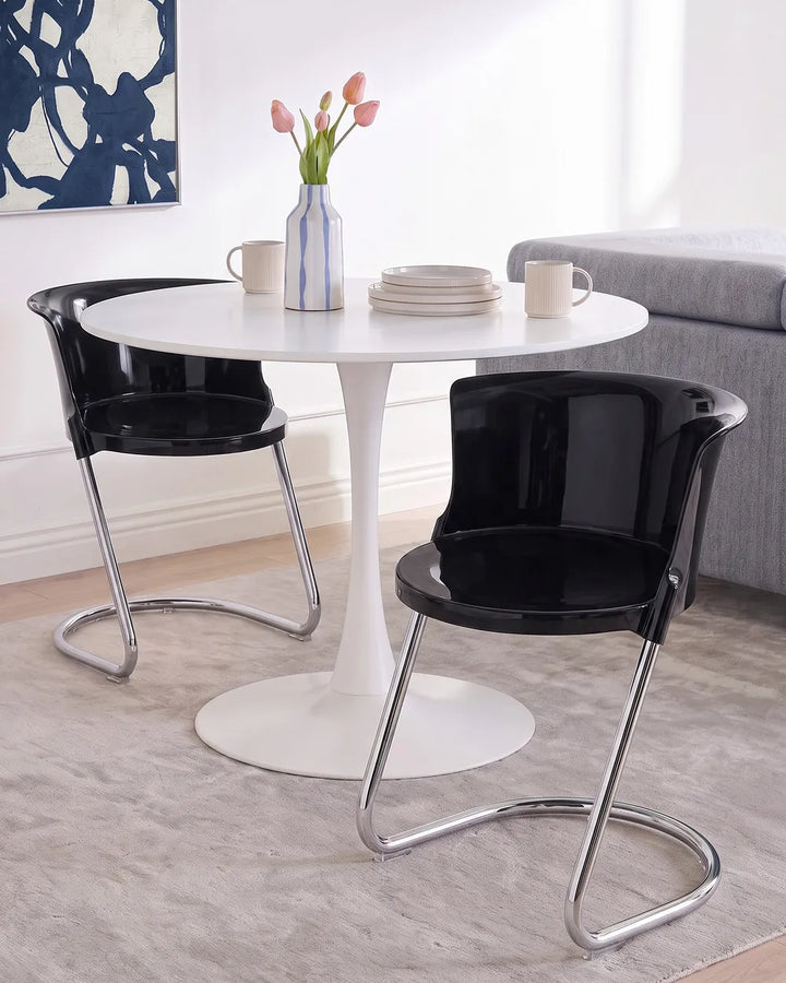 A modern dining setup featuring a round white pedestal table with two black curved-back chairs that have chrome metal legs, set on a light gray rug in a bright room.