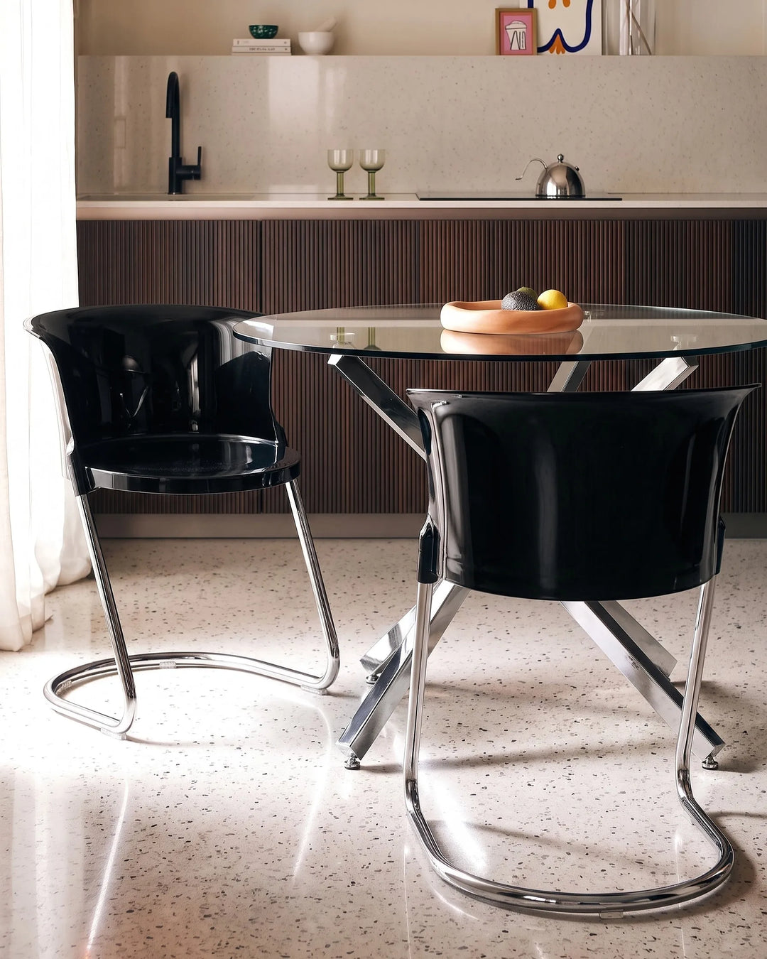 A modern dining setup featuring two glossy black chairs with chrome tubular steel legs around a round glass-top table, set in a contemporary kitchen with dark wood cabinets and a speckled terrazzo floor.