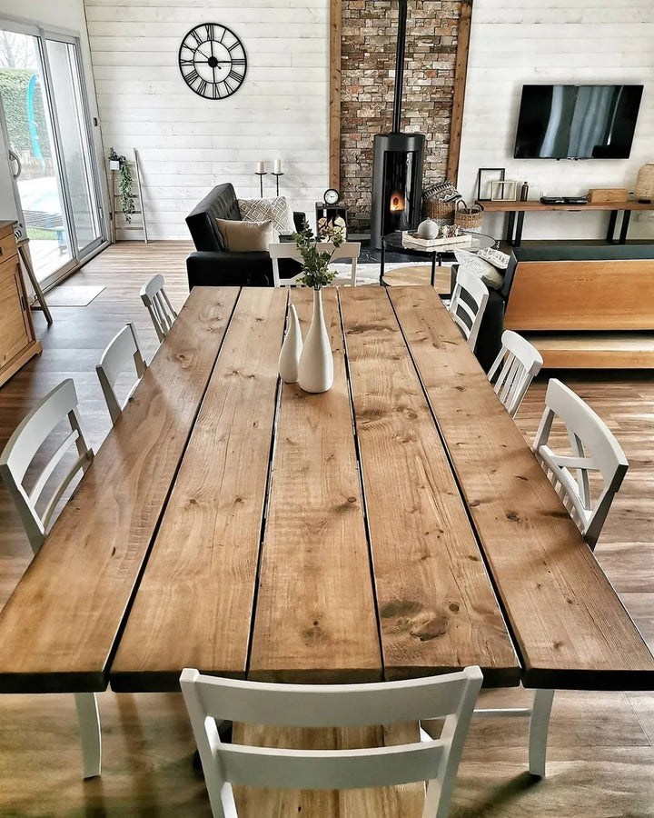A rustic wooden dining table with six white chairs, styled in a bright, modern farmhouse dining room with a brick fireplace and white shiplap walls.