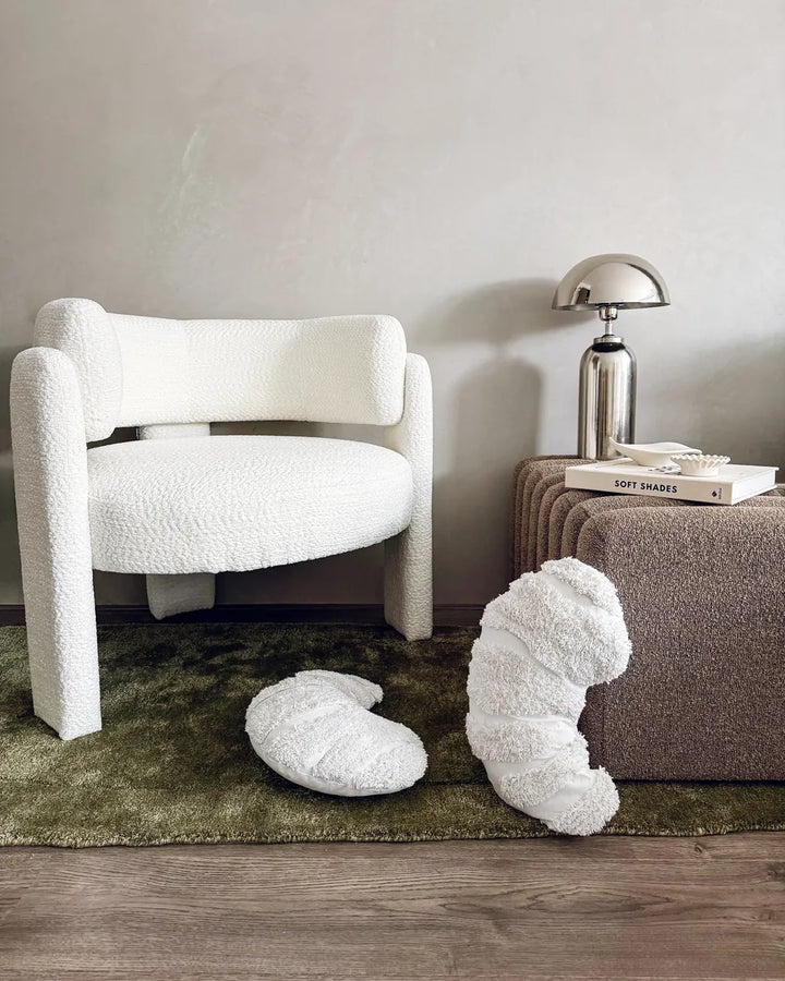 Modern white armchair with decorative pillows on a green rug in a room with a gray wall and brown side table.