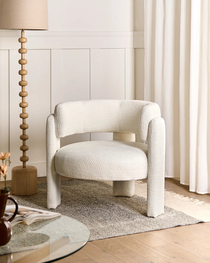 A modern round armchair upholstered in textured cream bouclé fabric, styled in a bright, neutral-toned living room next to a wooden floor lamp and a gray rug.