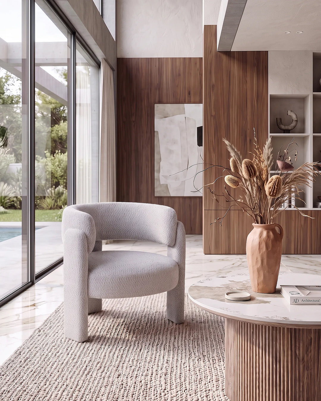 A modern light gray upholstered accent chair in a minimalist, contemporary living room with wood paneling, a marble coffee table, and a textured vase of dried grasses.