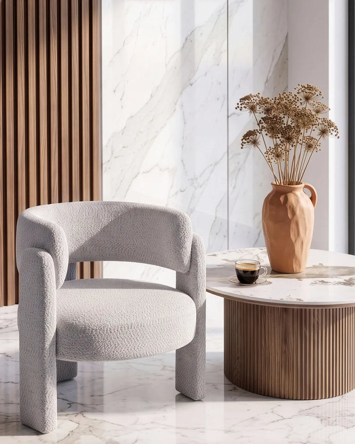 A modern upholstered accent chair in a textured light gray bouclé fabric, styled in a contemporary interior next to a marble-topped side table with a terracotta vase of dried flowers.