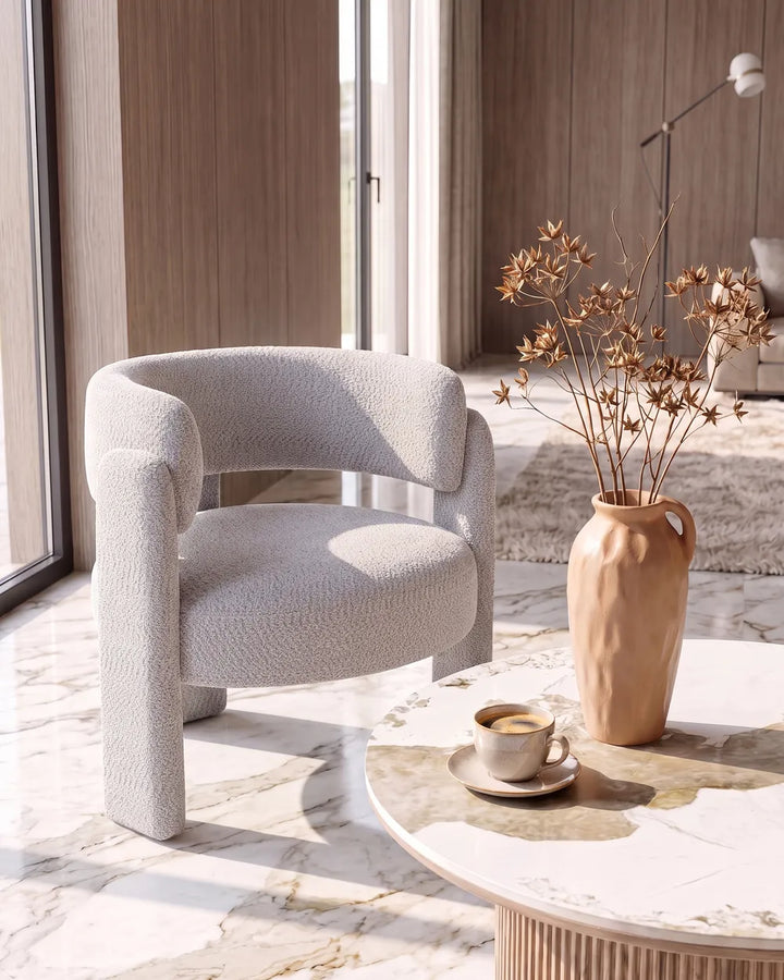 A modern curved armchair upholstered in a textured light gray fabric, styled in a bright, contemporary living room with a marble coffee table, a beige ceramic vase with dried branches, and a cup of coffee.