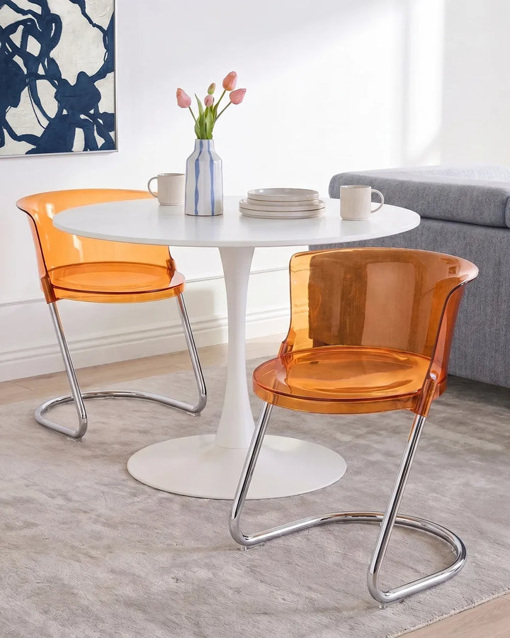Two modern translucent orange dining chairs with curved chrome metal legs, shown around a white round pedestal table in a bright dining room setting.