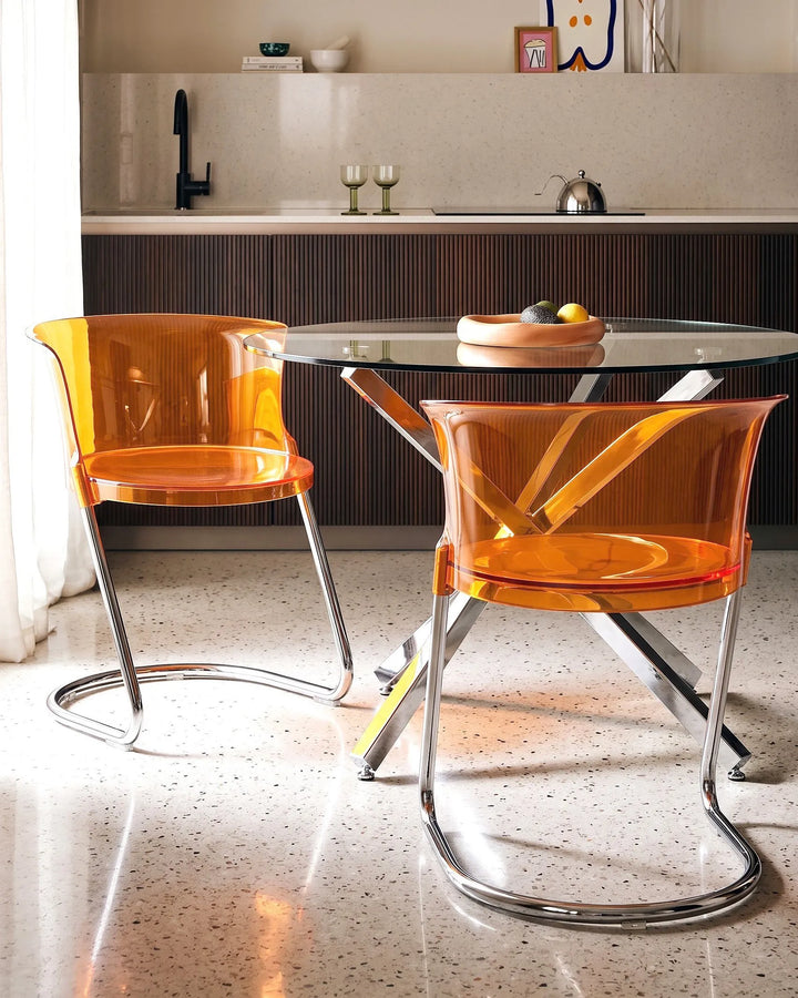 Two modern cantilever dining chairs with translucent orange plastic seats and chrome curved metal legs, positioned around a glass-top table in a contemporary kitchen setting.