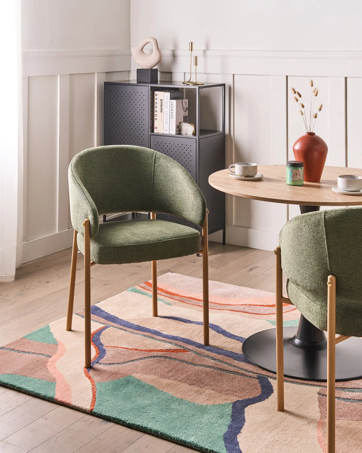 A styled interior scene showing two upholstered dining chairs with green fabric and light wood legs around a round wooden table on a patterned rug, with a dark storage cabinet in the background.