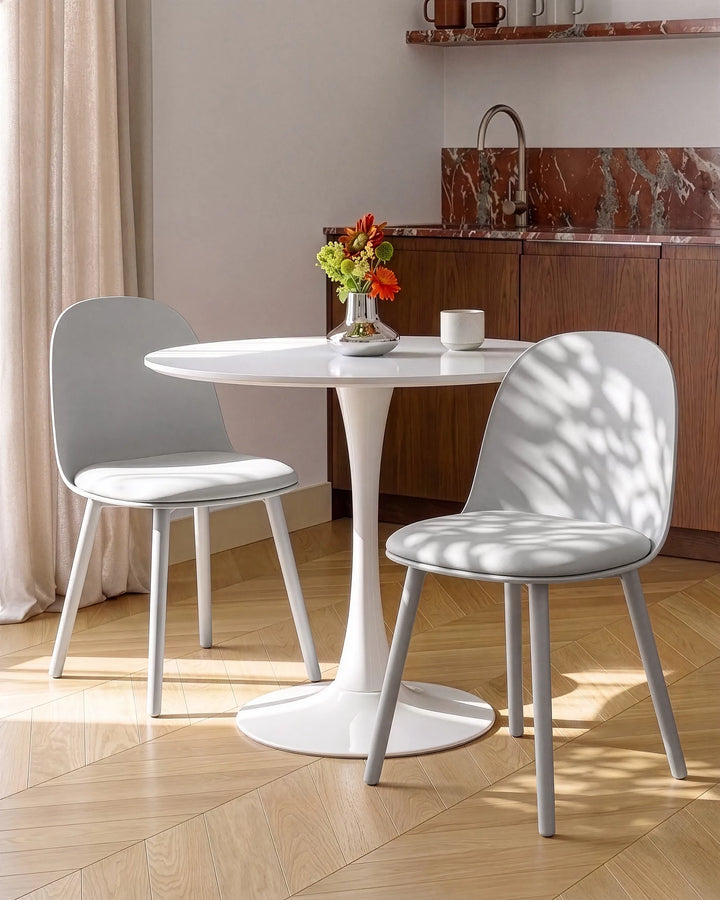 A modern dining set featuring a round white pedestal table and two light gray upholstered chairs, styled in a sunlit kitchen setting with wood flooring and a marble backsplash.