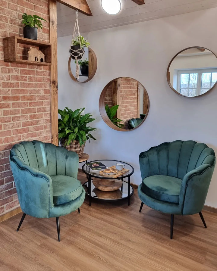 Two teal velvet shell-back armchairs in a styled room with a brick wall, wooden ceiling beams, round mirrors, and a small glass-top side table between them.