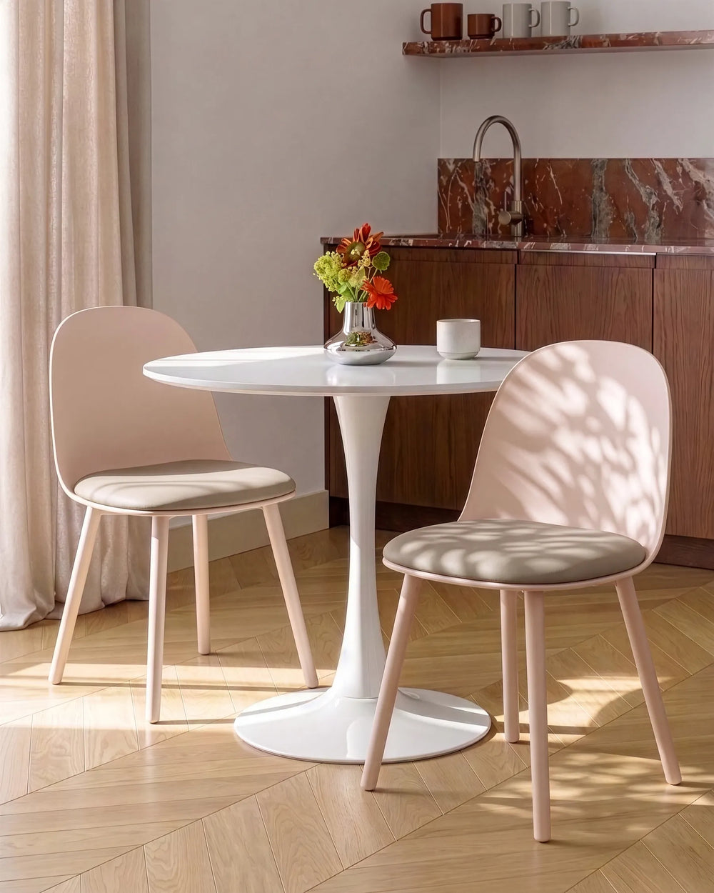A modern dining set featuring a round white pedestal table and two blush pink chairs with light wood legs, styled in a sunlit room with a marble kitchen background.