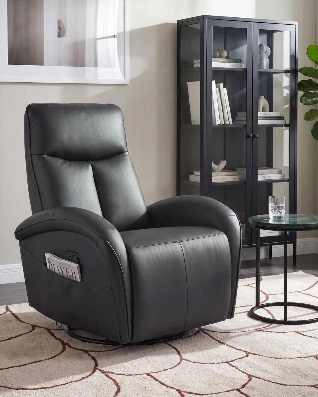 A black faux leather recliner chair with a headrest pillow and side control panel, shown in a styled living room setting next to a glass cabinet and side table.