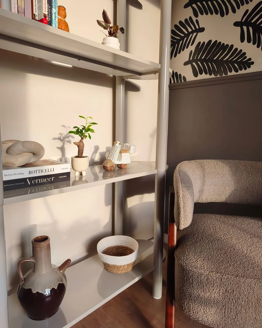 A modern white floating bookshelf with a metal frame in a styled room corner, holding books, a small potted plant, a ceramic vase, and a woven basket, next to a textured beige armchair.