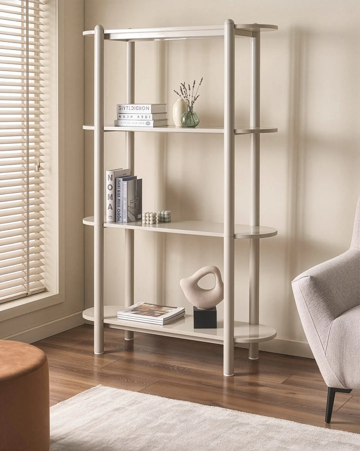 A tall, narrow open bookcase with four shelves in a light beige or off-white finish, styled with books, a small vase, and a sculptural object in a living room setting with wood floors and a chair.