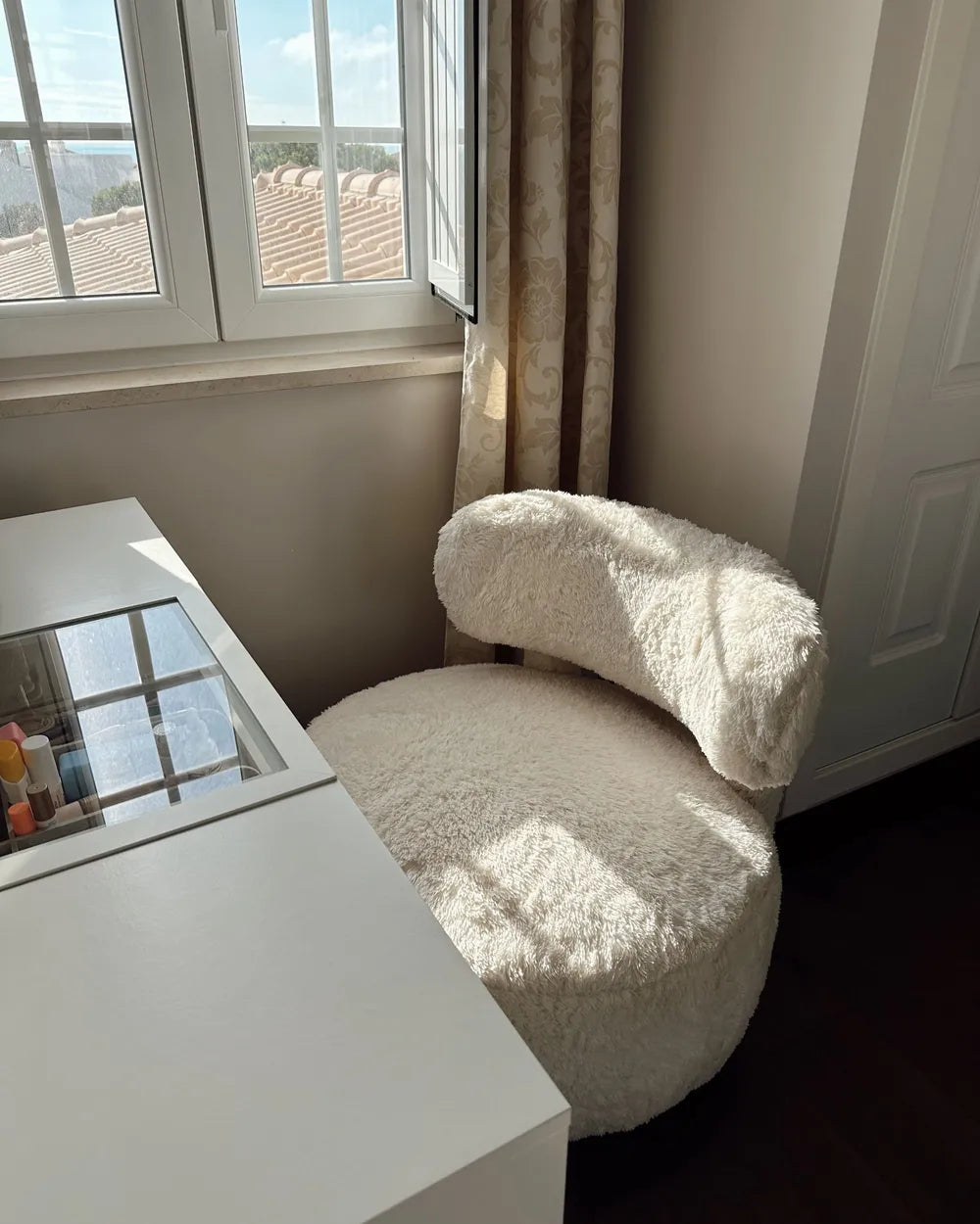 A round, fluffy cream-colored accent chair positioned next to a white vanity table in a sunlit room with a window and curtains.