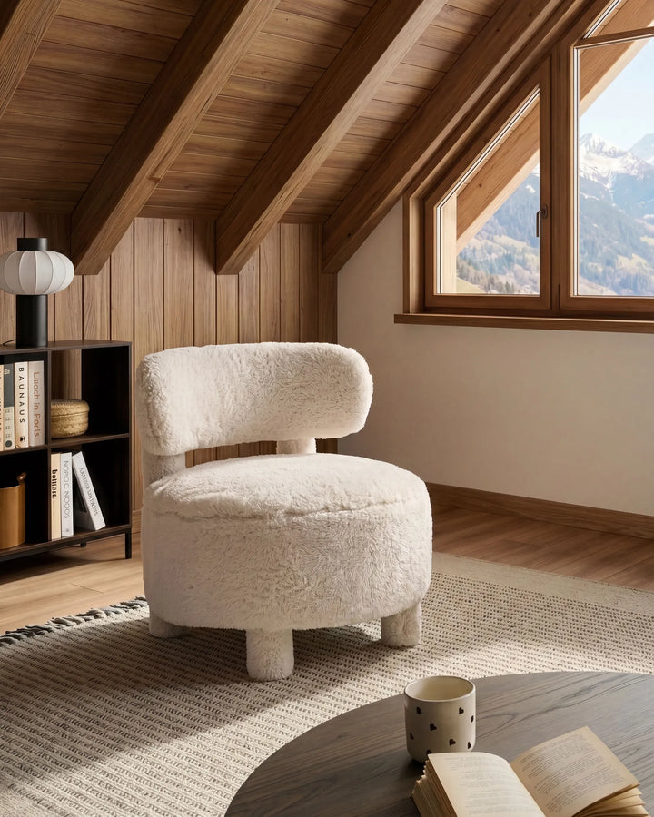A round, low-profile armchair upholstered in a fluffy cream bouclé fabric, positioned in a cozy attic room with wooden walls, a window looking out to mountains, and a wooden table in the foreground.