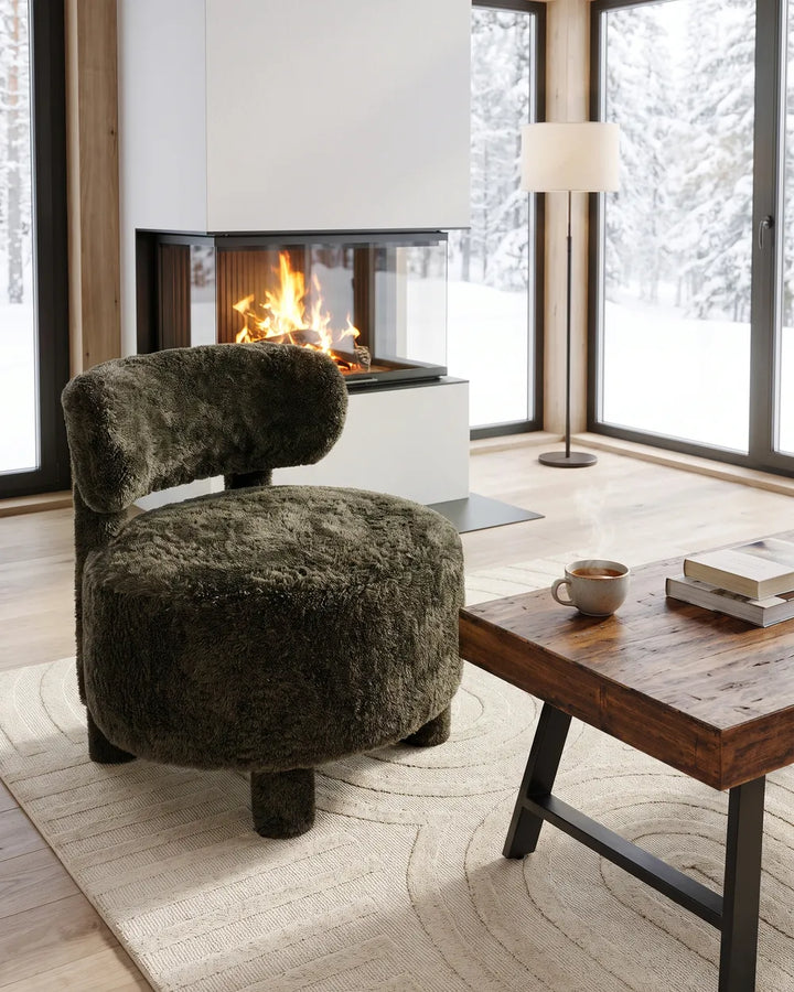 A round, low-profile armchair upholstered in dark olive green shaggy faux fur, positioned in a modern living room setting with a fireplace, large windows showing a snowy outdoor view, a wooden coffee table, and a beige rug.