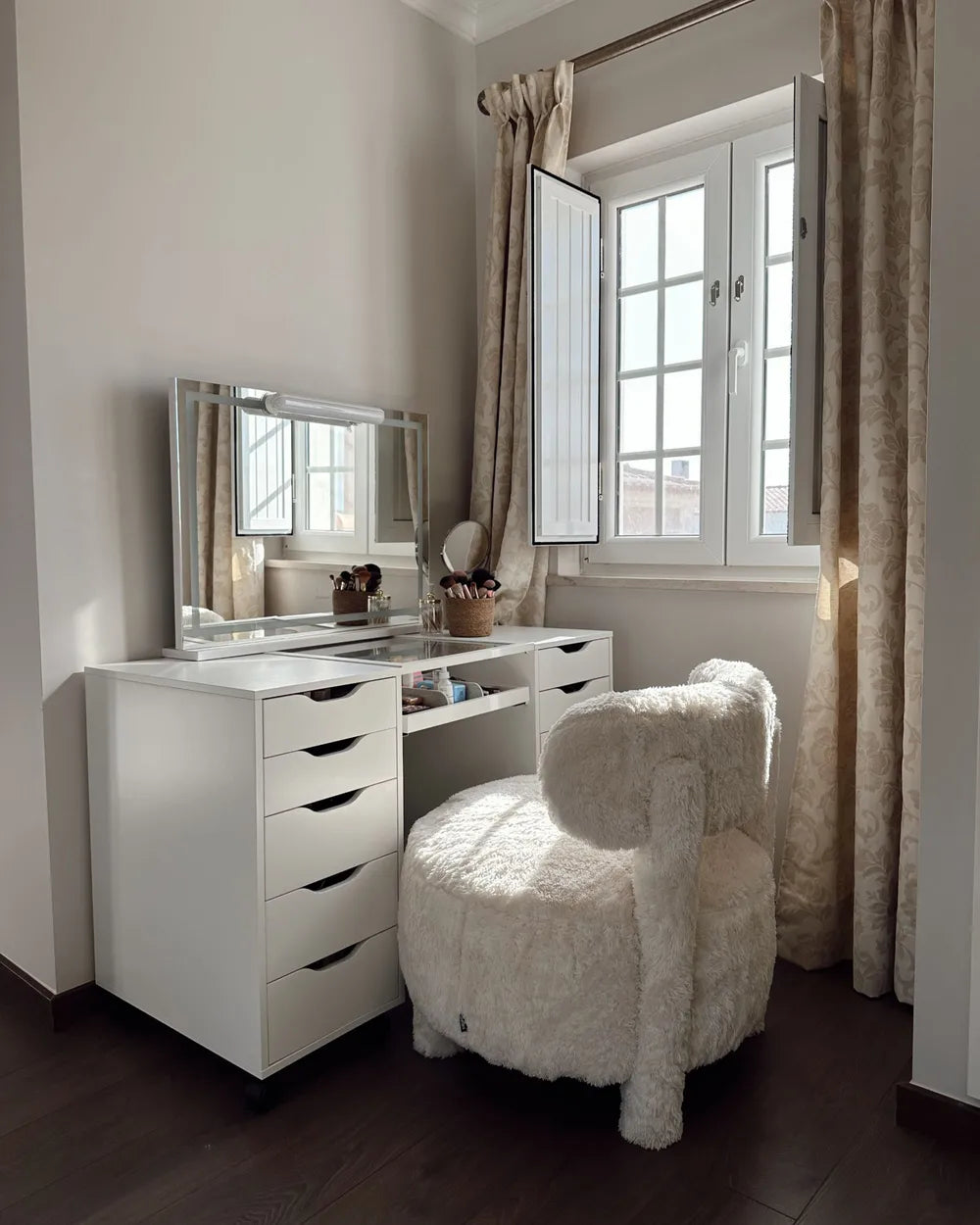 A white vanity desk with an attached rectangular mirror and six drawers, styled in a bright room next to a window with beige curtains. A fluffy cream-colored armchair sits in front of the desk.
