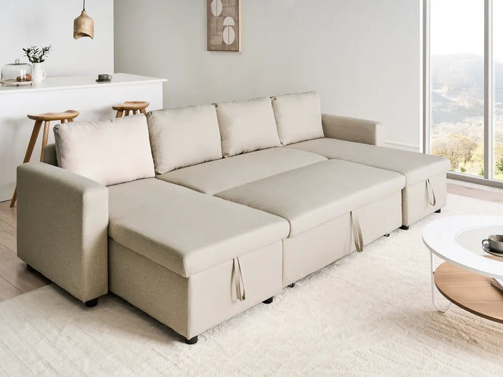 A light beige fabric sectional sofa with a chaise lounge on the left, styled in a bright, modern living room with a white coffee table and a large window showing a scenic view.