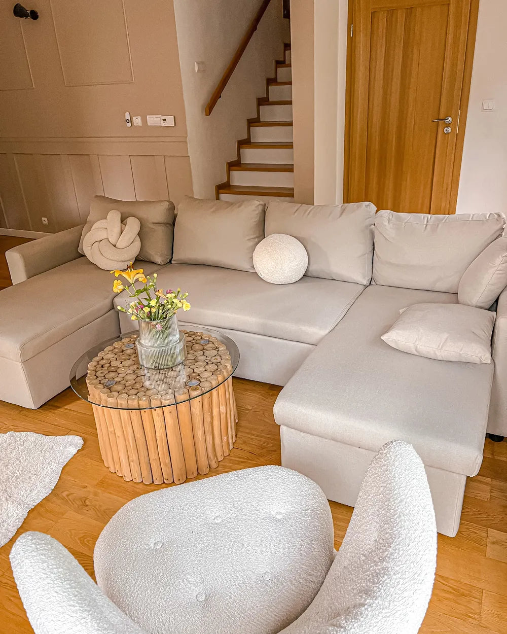 A light beige sectional sofa in a modern living room setting, styled with neutral throw pillows, a round white cushion, and a small wooden coffee table with a glass top.