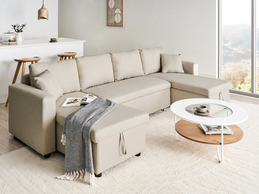 A light beige fabric sectional sofa with a chaise lounge, styled in a bright, modern living room with a gray throw blanket, a round coffee table, and a large window overlooking a scenic landscape.
