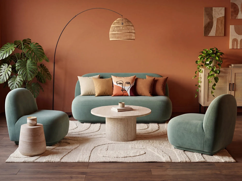 A styled living room scene featuring a teal-green velvet sofa with matching accent pillows, two matching teal-green armchairs, a round white coffee table, a beige area rug, a black floor lamp, and a terracotta-colored wall.